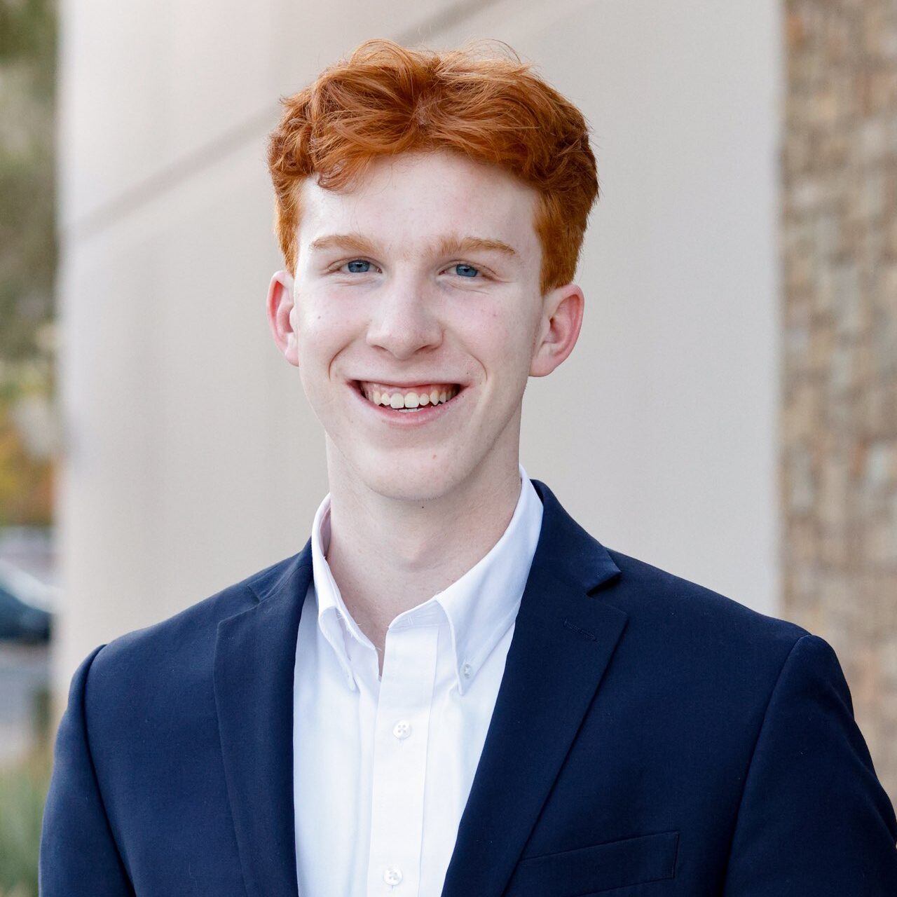 A person with red hair and a suit smiles outdoors.