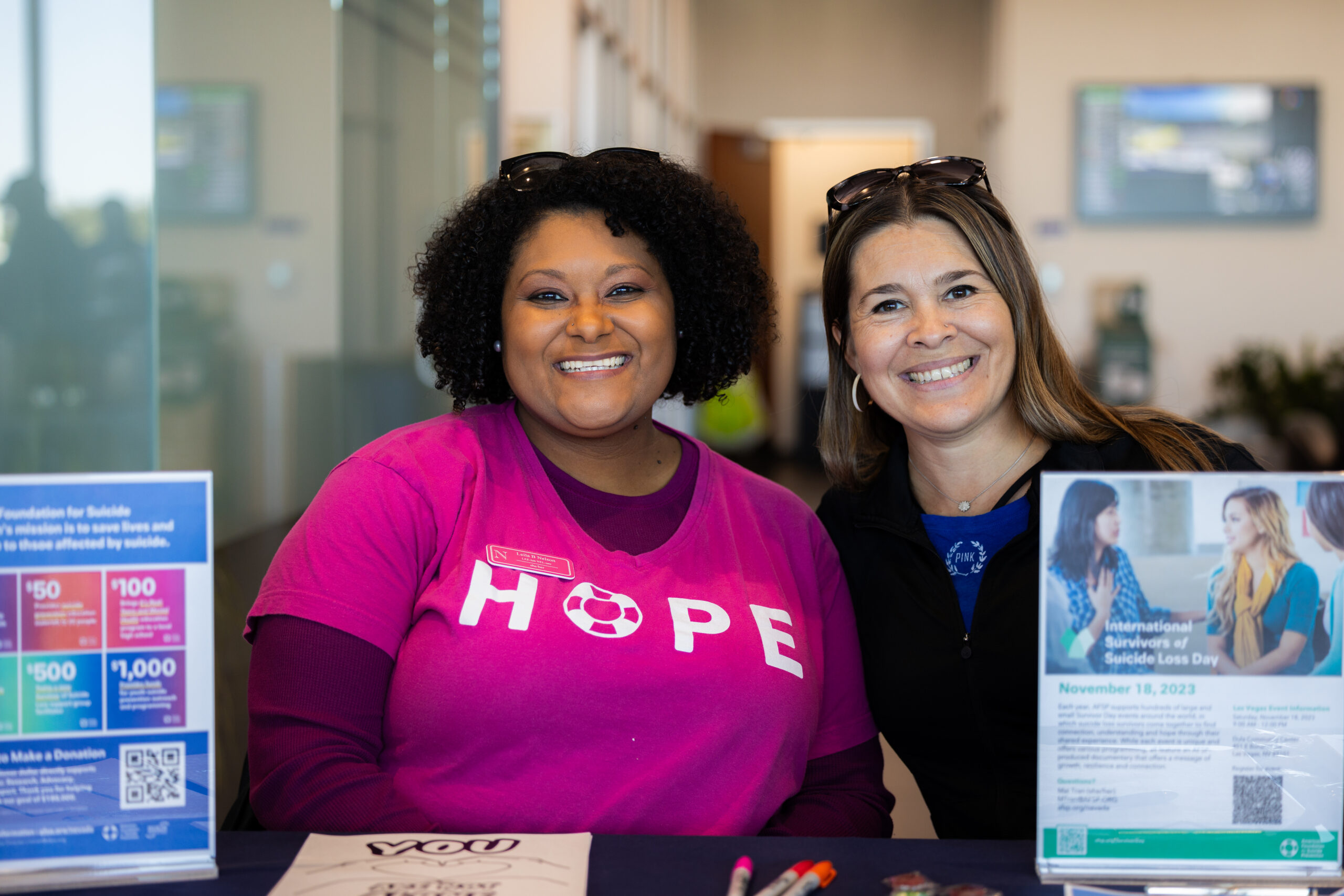 Two women smiling at a table, promoting suicide prevention awareness with informational materials.