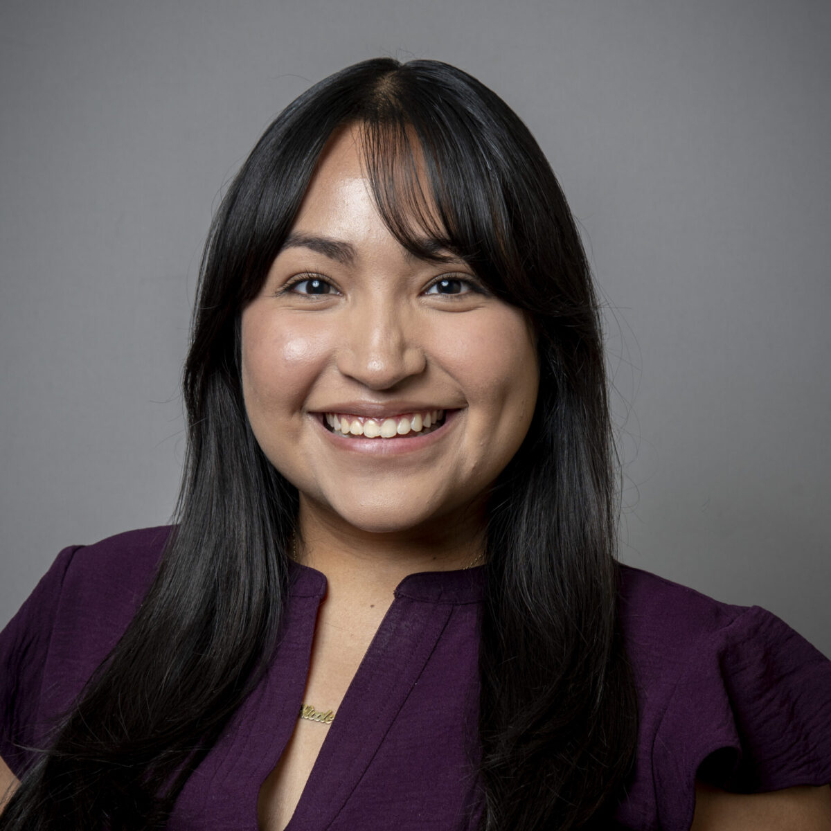 Smiling woman with long dark hair wearing a purple blouse, posed in front of a plain gray background.