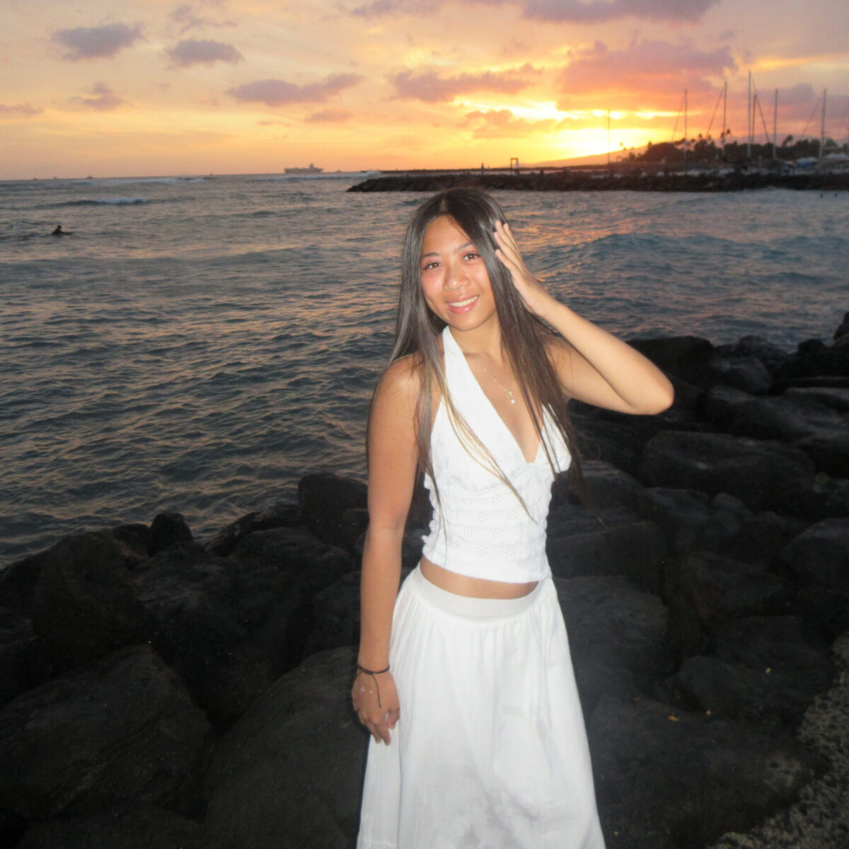 Woman in white dress stands on rocks by the ocean at sunset, with boats and clouds in the background.