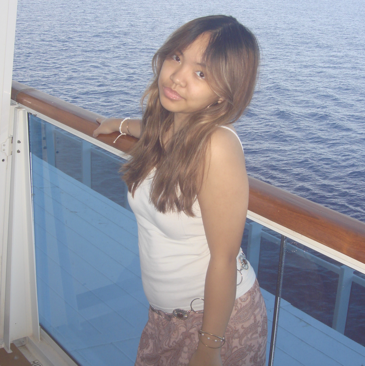 A young woman stands by a ship railing, with the ocean in the background on a bright day.