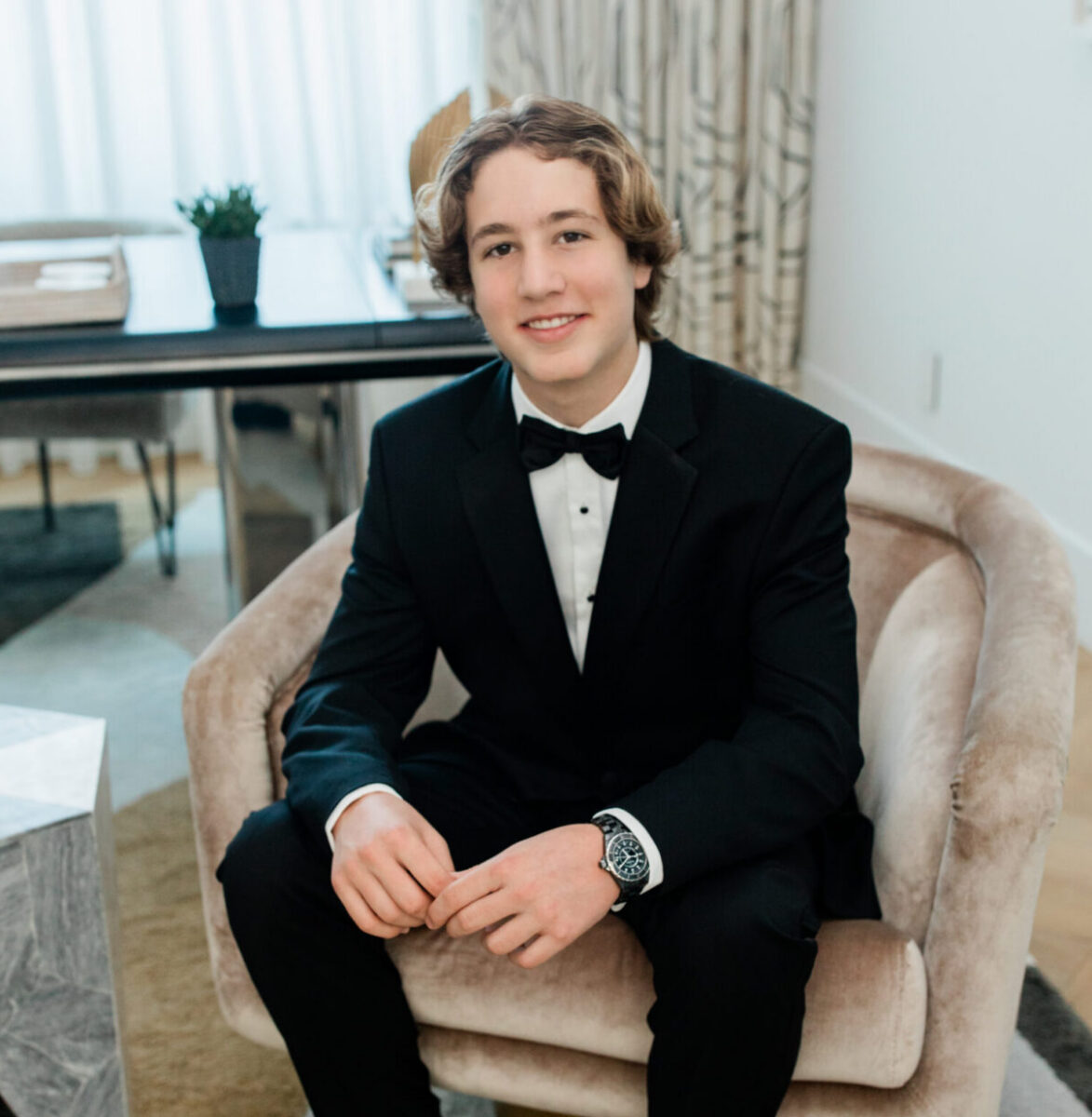 Young man in a black tuxedo sits smiling in a modern living room on a beige chair.